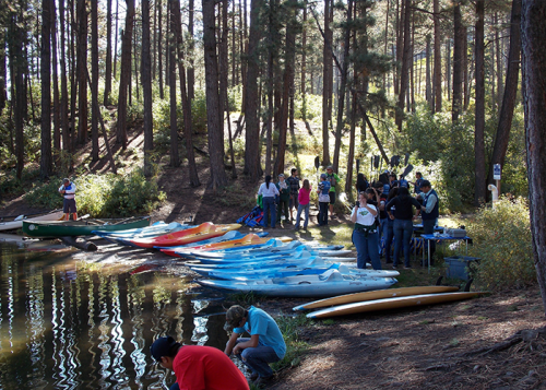 A group of people by a calm lake, with colorful kayaks lined up on the shore and trees in the background.