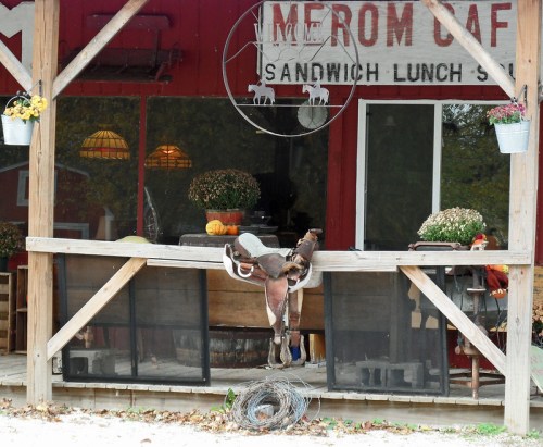 A rustic café with a wooden porch, featuring a saddle, potted flowers, and a sign for "Merom Café."