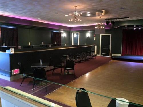 A dimly lit performance space with empty chairs, a small stage, and purple lighting along the ceiling.