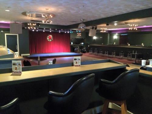 A dimly lit performance venue with a stage, red curtains, and bar seating, ready for an event.