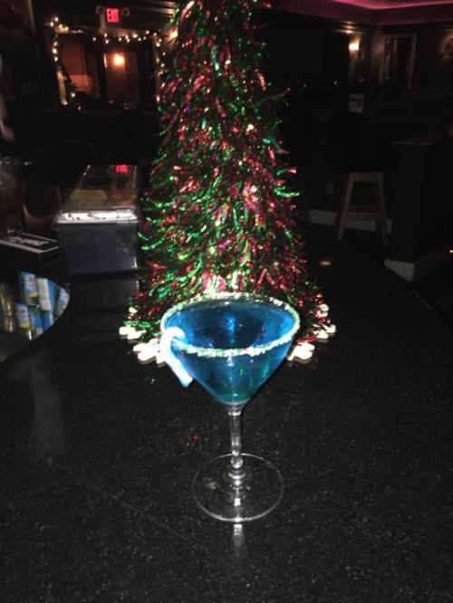 A blue cocktail in a glass with a festive tree in the background, decorated with red and green tinsel.