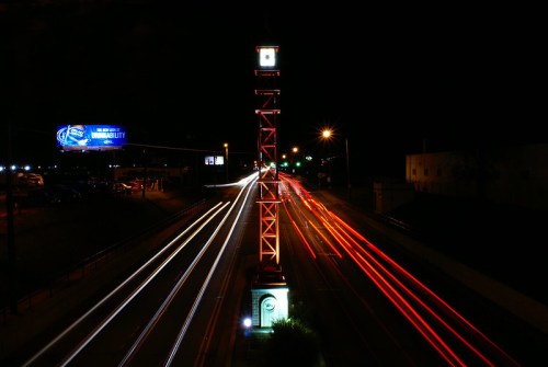 A night scene featuring a clock tower beside a busy road, with light trails from passing vehicles and a billboard in the background.