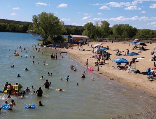 A busy beach scene with people swimming, lounging under umbrellas, and enjoying the water on a sunny day.