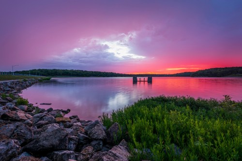 A serene lake at sunset, with vibrant purple and pink skies reflecting on the water and rocky shores in the foreground.