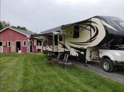 A parked RV next to a red barn, with lawn chairs set up on green grass.