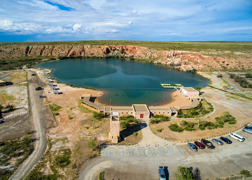 Aerial view of a large pond surrounded by rocky cliffs, with a building and parking area nearby. People are swimming.