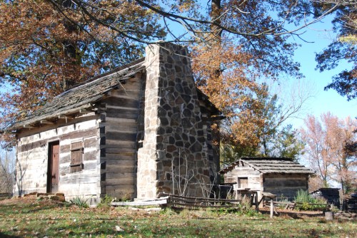 A rustic log cabin with a stone chimney, surrounded by trees with autumn leaves. A smaller shed is nearby.