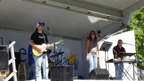 A band performs on stage with a guitarist, a female vocalist, and a keyboardist, surrounded by musical equipment.