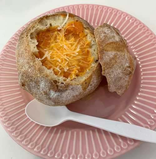 A bread bowl filled with orange soup and topped with shredded cheese, served on a pink plate with a white spoon.