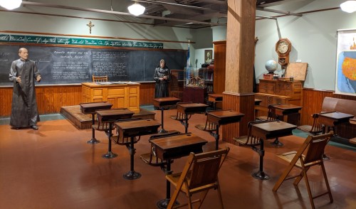 A vintage classroom with wooden desks, a chalkboard, and two figures, one at the front and one by the side.
