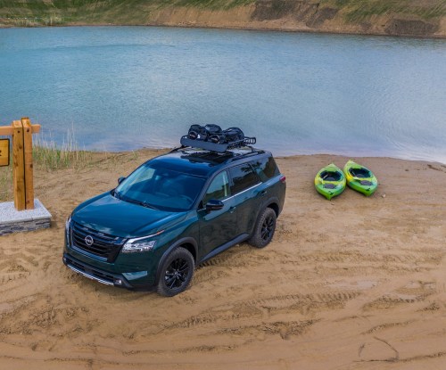 A green SUV parked on sandy shore near a calm lake, with two green kayaks nearby and a wooden sign in the background.