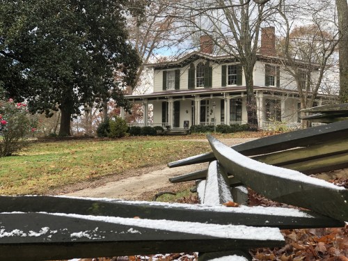 A historic white house with a porch, surrounded by trees and a dusting of snow on the ground and nearby benches.