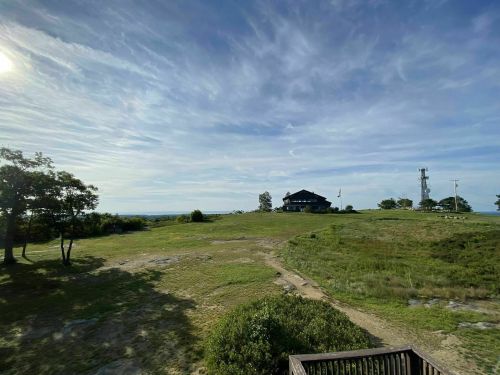 A grassy landscape with a distant house and communication tower under a blue sky with wispy clouds.