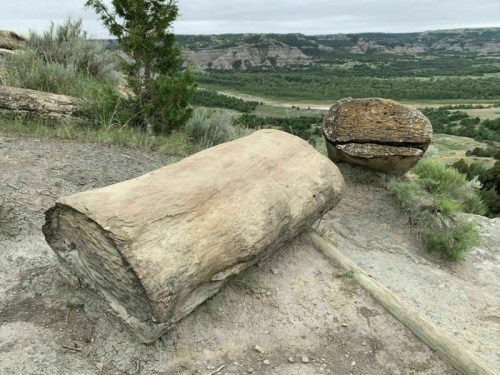 Two large, weathered rocks sit on a hillside overlooking a green valley and distant hills under a cloudy sky.