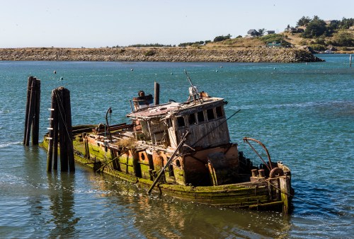 A weathered, abandoned boat partially submerged in calm waters, surrounded by a scenic landscape and distant shoreline.