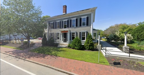 A two-story building with white siding, black shutters, and a brick walkway, surrounded by greenery on a sunny day.