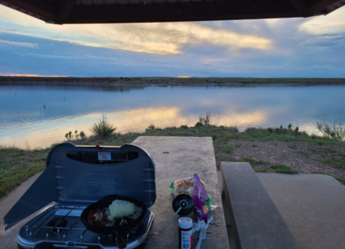 A camping stove with food cooking beside a calm lake at sunset, with clouds reflecting on the water.