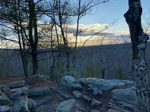 A serene landscape featuring rocky terrain, trees, and a sunset sky over a distant forest.