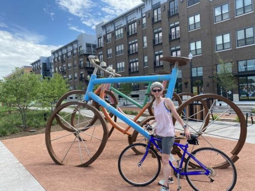 A person stands next to a large, colorful bicycle sculpture in an urban park, holding a smaller blue bicycle.