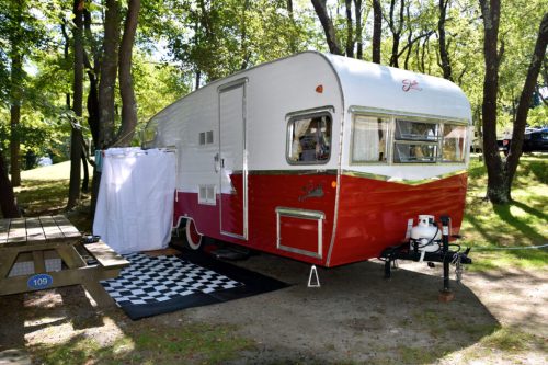 A vintage red and white camper trailer parked in a wooded area, with a picnic table and checkered mat nearby.