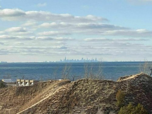 A distant view of a city skyline across a body of water, with sandy hills in the foreground and cloudy skies.