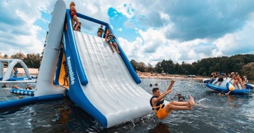 A group of people enjoying an inflatable water slide at a lake, with others playing nearby under a cloudy sky.