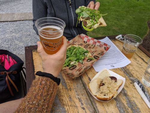 A hand holds a beer while another hand lifts a sandwich topped with greens, with a plate of food on the table.