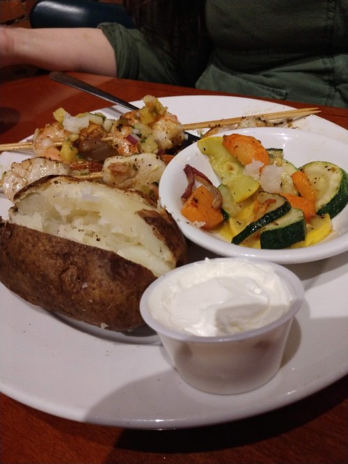 A plate with a baked potato, grilled shrimp, and a side of mixed sautéed vegetables. A small cup of sour cream is included.