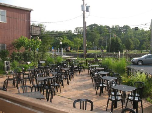 Outdoor seating area with empty black tables and chairs, surrounded by greenery and a view of a park in the background.