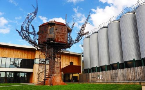 A large, artistic tree structure made of metal stands next to silos and a modern building under a blue sky.