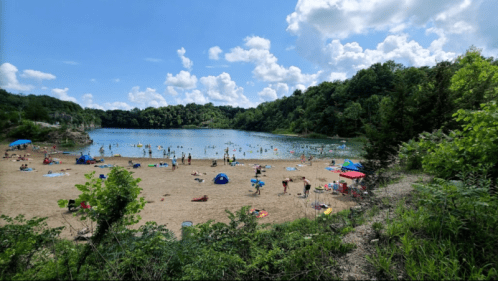 A sandy beach by a lake, with people swimming and relaxing under a blue sky with fluffy clouds and green trees.