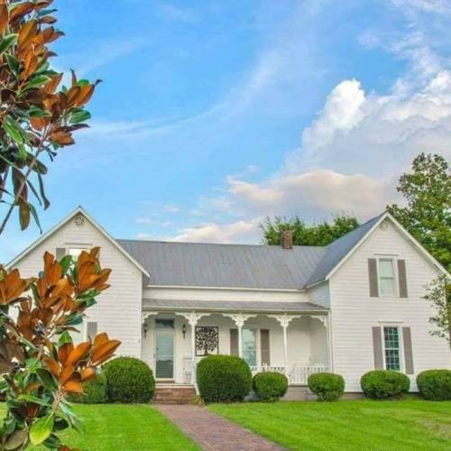 A charming white house with a gray roof, surrounded by green grass and shrubs, under a blue sky with fluffy clouds.