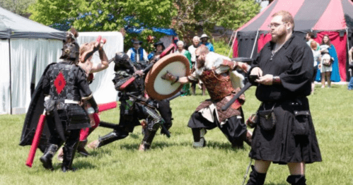 A group of people in medieval costumes engage in a staged battle on a grassy field during a festival.