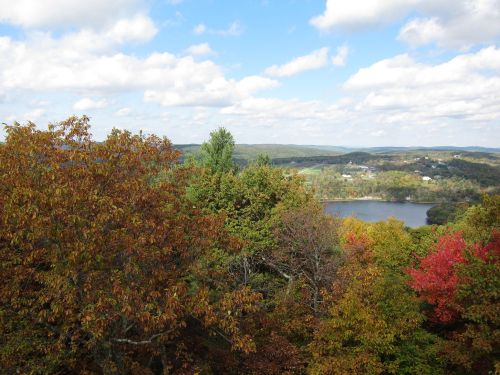 A scenic view of a lake surrounded by trees with autumn foliage under a partly cloudy sky.