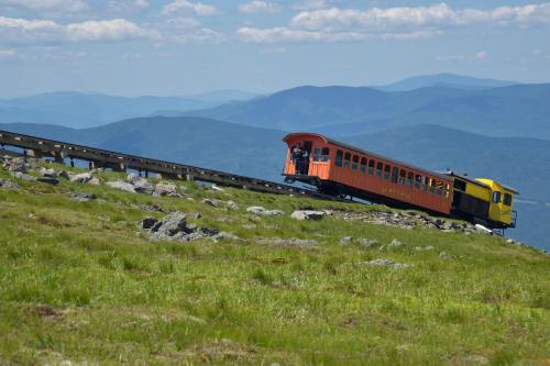 A colorful train climbs a steep track on a mountain, surrounded by green grass and distant blue hills under a cloudy sky.