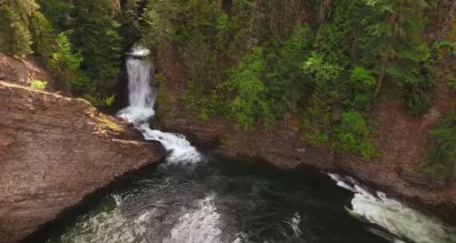 Aerial view of a serene waterfall cascading into a dark pool, surrounded by lush green trees and rocky cliffs.