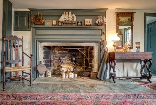 Cozy living room with a stone fireplace, wooden chair, decorative ships, and a vintage table on a patterned rug.