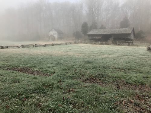 A misty landscape featuring two rustic barns surrounded by a frosty field and trees in the background.