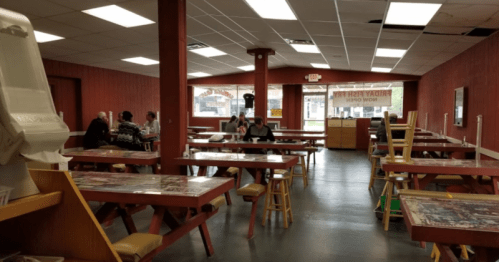 A spacious, empty dining area with wooden tables and stools, featuring a few patrons and a counter in the background.