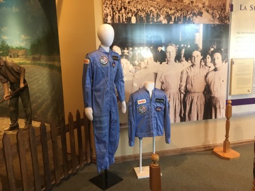 Two blue astronaut suits displayed on mannequins, with a historical photo of a crowd in the background.