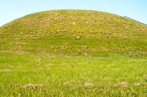 A grassy hill with a smooth, rounded top, surrounded by a lush green field under a clear blue sky.