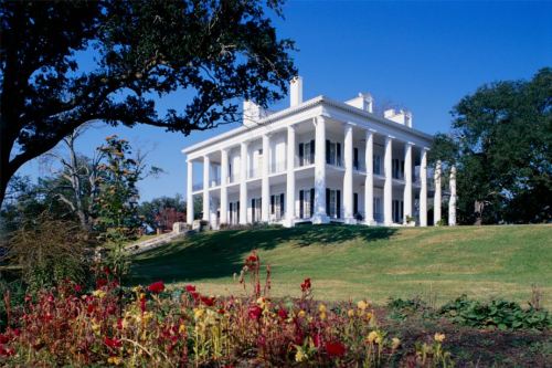 A grand white mansion with columns, set on a grassy hill, surrounded by colorful flowers under a clear blue sky.