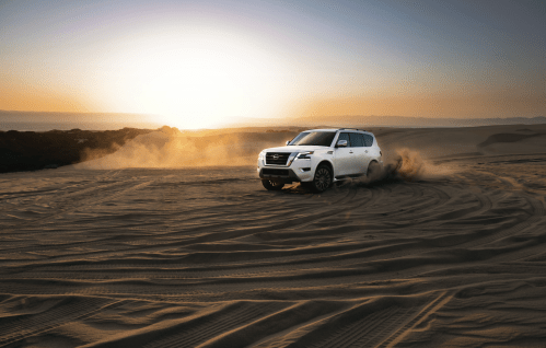 A white SUV driving through sandy terrain at sunset, kicking up dust against a backdrop of a glowing horizon.