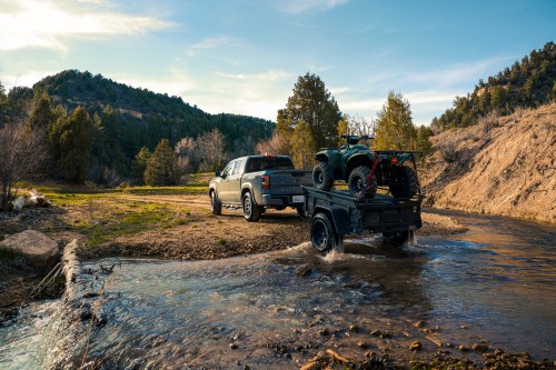 A truck towing a trailer with an ATV crosses a shallow stream in a scenic outdoor setting.