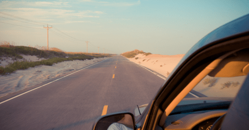 A view from a car window of a long, empty road lined with sand and grass under a clear sky.