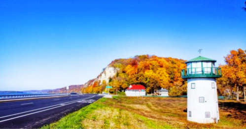 A scenic view of a lighthouse by a road, with vibrant autumn foliage on a hillside and a clear blue sky.