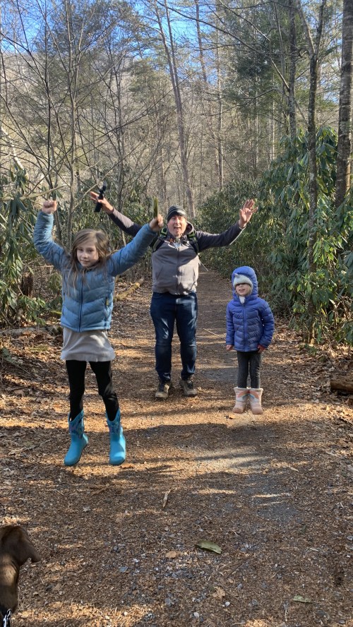 A man and two children joyfully jump on a forest trail, surrounded by trees and greenery.