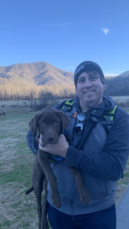 A man in a beanie holds a brown puppy, with mountains and a field in the background.