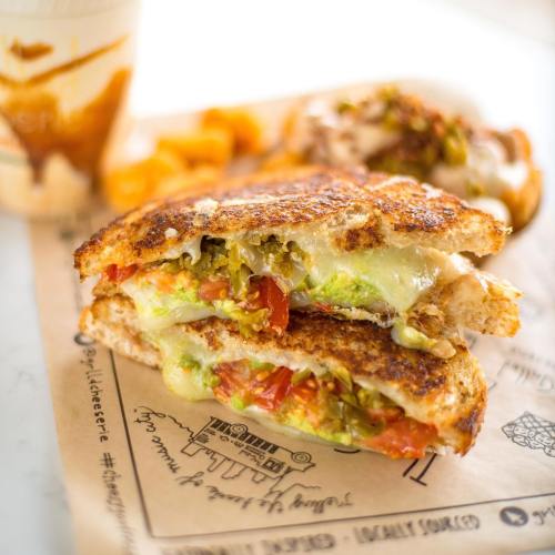 A grilled cheese sandwich with melted cheese, tomatoes, and green toppings, served with a drink and orange slices.