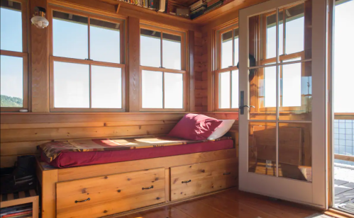 Cozy wooden interior with a bed by the window, sunlight streaming in, and a door leading outside.